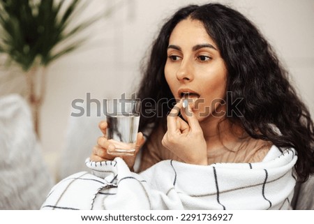 Beautiful young woman holding a pill and water. In the foreground a glass of water and a pill. In the background is an Arab woman looking at a pill. The girl thought about taking medicine.