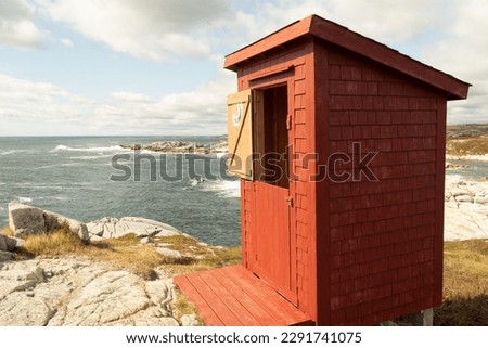Red wooden outhouse,  closeup, with 2 part door, a slanted roof and shingle siding, on a rocky shore, looking over the ocean. The upper half of the door is open with a moon, bathroom, sign