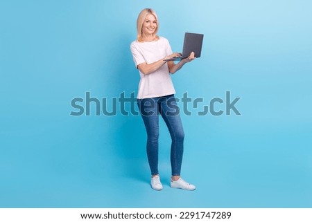 Full size photo of positive nice senior woman white t-shirt jeans sneakers writing letter on laptop isolated on blue color background