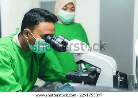 Southeast Asian Scientist Looking Under Microscope, With Medical Assistant, Wearing Green Protective Clothing. Hospital Medical Development Laboratory. Medicine, Biotechnology, Microbiology, and Drugs Royalty-Free Stock Photo #2291755097