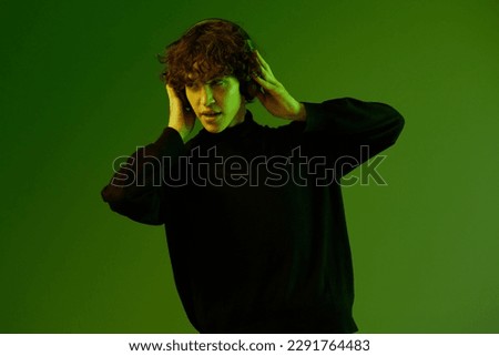 Male teenager in headphones listening to music, dancing and singing with his eyes closed, DJ happiness and smile, hipster lifestyle, portrait green background mixed neon light, copy space