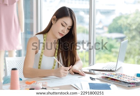 Millennial Asian young beautiful professional female dressmaker designer seamstress using pencil drawing sketching designing trendy style outfit on working desk in tailor workshop studio office.