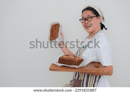 A middle-aged woman wearing an apron over white t-shirt is holding cakes in her hands 