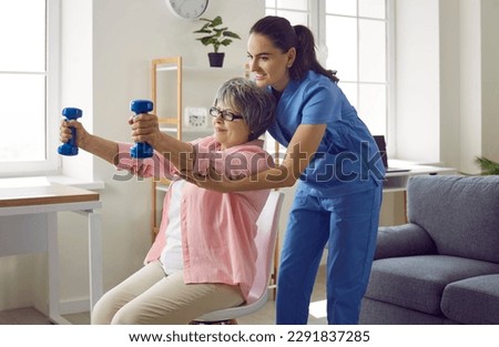 Young nurse or physiotherapist in scrubs helping a happy retired old woman do fitness exercises with light weight dumbbells at home. Concept of physiotherapy for seniors Royalty-Free Stock Photo #2291837285