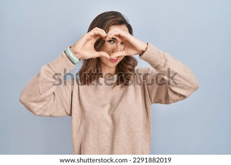 Young woman standing over isolated background doing heart shape with hand and fingers smiling looking through sign 