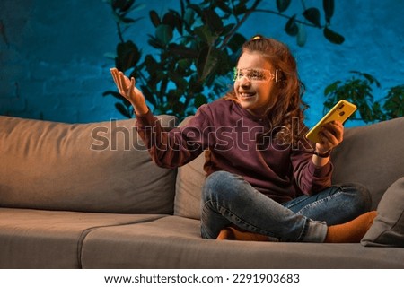 Teen girl in futuristic luminous glasses. Teenager sits on sofa and surfs social media and uses mobile app on smartphone in blue neon light living room at home. Gadget addiction concept