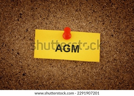 A yellow paper note with the words AGM (Annual General Meeting) on it pinned to cork board. Close up. Royalty-Free Stock Photo #2291907201