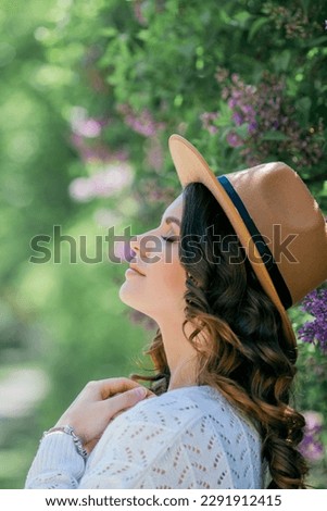 Portrait of beautiful young dark-haired woman with blue eyes in hat near the blooming. Happy model with curly hair.