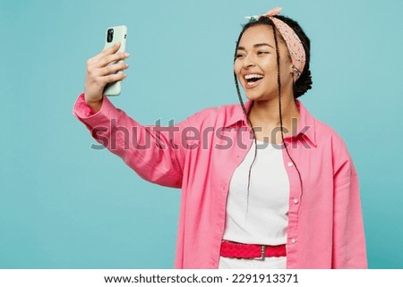 Young woman of African American ethnicity wear pink shirt white t-shirt doing selfie shot on mobile cell phone post photo on social network isolated on plain pastel light blue cyan background studio
