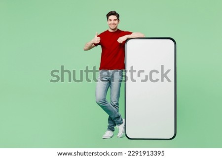 Full body happy fun young man he wears red t-shirt casual clothes big huge blank screen mobile cell phone smartphone with workspace area show thumb up isolated on plain pastel light green background