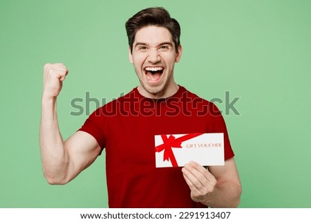 Young surprised happy man he wearing red t-shirt casual clothes hold gift certificate coupon voucher card for store do winner gesture isolated on plain pastel light green background studio portrait