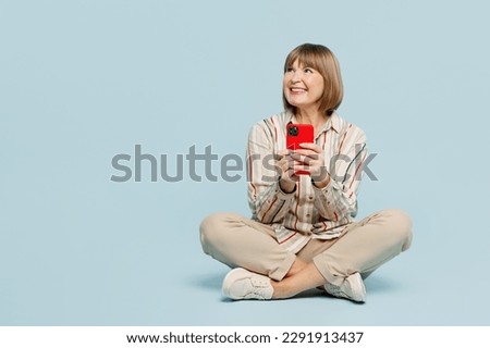 Full body elderly woman 50s years old wear shirt sitting hold in hand use mobile cell phone look aside on area isolated on plain pastel light blue color background studio portrait. Lifestyle concept
