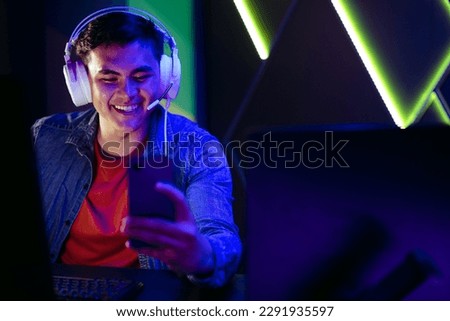 Happy young gamer watching and streaming live game broadcasts on his smartphone. Wearing his headset, he connects and participates in virtual gaming communities using an online streaming app. Royalty-Free Stock Photo #2291935597