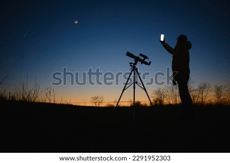 Astronomer looking at the starry skies with planets, falling stars and Moon eclipse with a telescope while using modern smartphone.