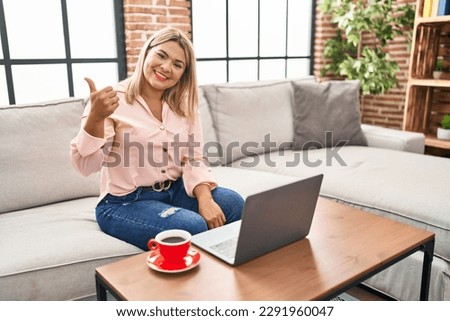 Young hispanic woman using laptop sitting on the sofa at home doing happy thumbs up gesture with hand. approving expression looking at the camera showing success. 