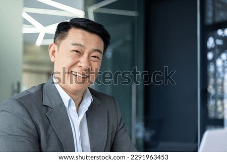 Close up portrait of successful asian financier investor, adult boss smiling and looking at camera, man working inside office at workplace with laptop.