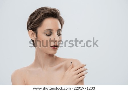 portrait of young woman with bright makeup and short hair touching bare shoulder and looking at camera isolated on grey