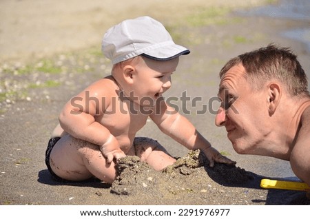 A small tanned boy in a white cap sits on the shore of a sandy beach and smiles at his dad. Close-up portrait. Dad and baby on the sea, family vacation concept Royalty-Free Stock Photo #2291976977
