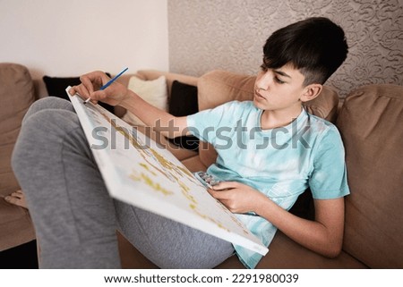 Young male teenager painting picture by numbers on canvas in living room at home. Hobby and leisure concept.