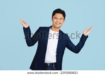 Surprised handsome Asian man in semi formal clothes smiling and opening hands in light blue isolated studio background Royalty-Free Stock Photo #2292000821
