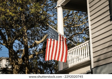 A patriotic display of an American Flag hanging on the side of a home in direct sunlight.