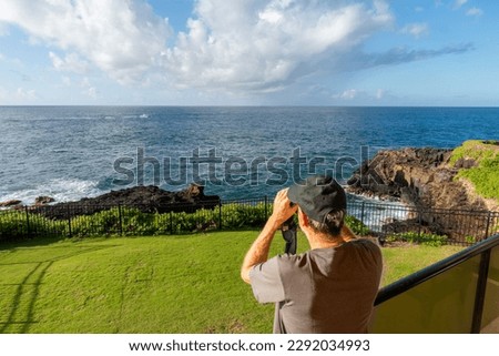 Mature Caucasian man watching a boat far on the ocean with binoculars from a balcony, lanai, with grass, iron fence and rocky cliff, Makahuena Point, Kauai, Hawaii