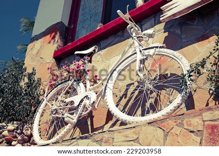 White vintage bicycle with red flowers stands near old stone wall. Retro toned effect, Instagram style photo filter