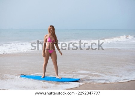 Young beautiful blonde surfer woman in pink bikini standing on blue surfboard. The girl enjoys her holidays on the beach to practice her favourite sport. Travel and holiday concept