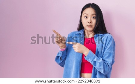 Young asian woman standing over pink background pointing aside worried and nervous with both hands, concerned and surprised expression 