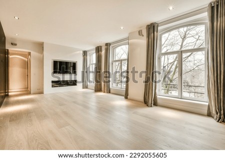 an empty living room with wood flooring and large windows looking out onto the trees that line the street outside Royalty-Free Stock Photo #2292055605