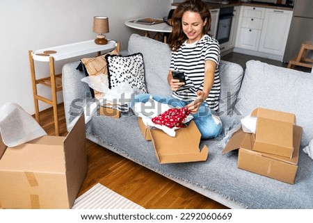 Happy woman sharing photos of her online purchase on her social media while sitting on sofa and unpacking cardboard boxes.