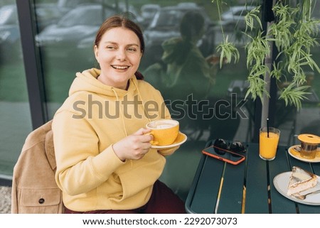 Happy Young woman enjoying a coffee, sitting on the cafe terrace on the modern city street. Person sitting at restaurant table and drinking cappuccino in ceramic yellow cup, Concept of easy breakfast