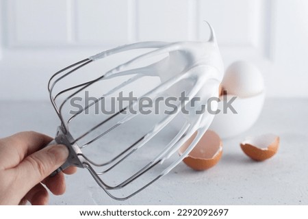 Whisked egg whites - whipped Italian meringue on a wire whisk and eggs on a gray background. copy space. Royalty-Free Stock Photo #2292092697