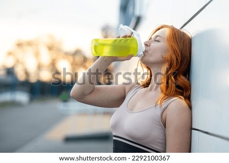 red-haired young woman leaning against the wall drinks water. after workout outdoors in summer