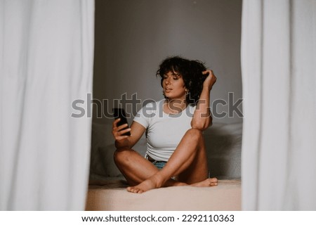 Portrait of a romantic woman sitting in bed with a smartphone and with headphones. Young lady browsing through dating apps, listening music, using social media, relaxing at home. Copy space. Royalty-Free Stock Photo #2292110363