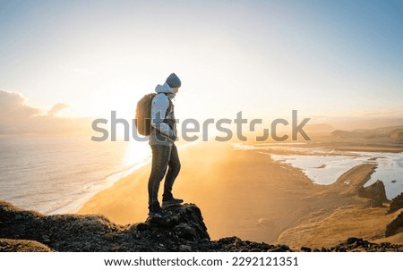 Lone traveler stands on a rocky cliff, taking in the stunning view. The warm glow of the sun's rays cast a golden light over the water. Inspiration and a sense of wanderlust. Royalty-Free Stock Photo #2292121351
