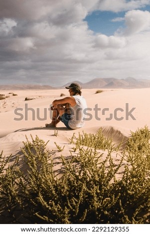 Young man with cap sitting on the desert sand of the dunes of Corralejo in Fuerteventura, looking at the horizon, in summer, during a vacation trip through the Canaries