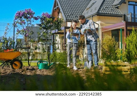 Caucasian father and pretty teen daughter planting tree together at house. Outdoor. Work in garden. Beautiful girl showing something on tablet computer to her dad in garden.