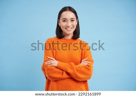 Young asian woman looks with confidence, cross arms on chest and smiles at camera, stands in orange sweatshirt, blue background.