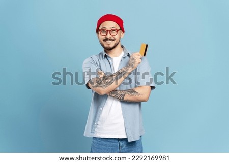 Successful smiling Asian man wearing red hat, stylish eyeglasses, holding credit card looking at camera isolated on blue background. Happy confident freelancer receive payment. Shopping, sales concept