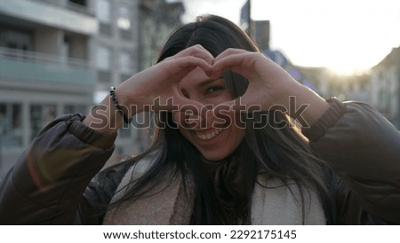 One happy young woman doing heart symbol with hands standing in city street during sunset time. Portrait face of a person makes heart shape gesture