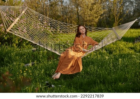 horizontal photo of a beautiful, red-haired woman lying in a hammock enjoying a rest in a long orange dress, on a warm summer day, smiling happily looking at the camera