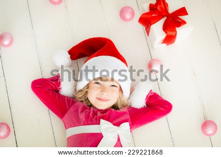 Happy child dreaming of Christmas gift. Funny kid dressed in Santa Claus hat. Baby lying on wood floor at home. Xmas holiday concept