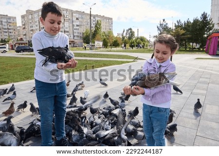 Two adorable kids, brother and sister feed pigeons in the park square. Spring. Lifestyle. The concept of kindness, care for animals. Family outing. A walk with benefit. Children, nature and animals.