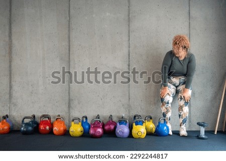 Exhausted young plus size woman in fitness clothes taking a break from exercising, feeling tired, resting during workout with weights, exhaling and looking at colorful kettlebells in gym. Copy space