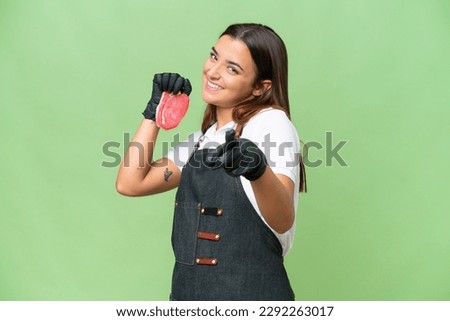 Butcher woman wearing an apron and serving fresh cut meat isolated on green chroma background pointing front with happy expression