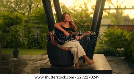 Beautiful Indian woman sitting on chair playing acoustic guitar and singing alone outside home. Happy Cheerful female enjoy sing some new sound on classical guitar Learning, practicing as hobby  Royalty-Free Stock Photo #2292270249
