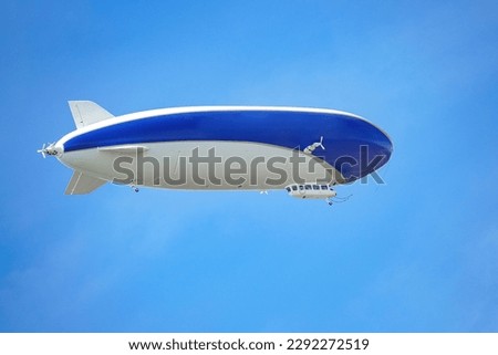 A blue and white airship with passengers on board flies against the blue sky.  Royalty-Free Stock Photo #2292272519