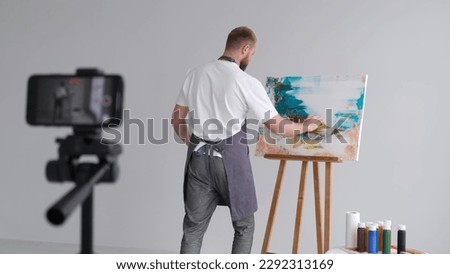An artist in a work coat records a video blog as he paints a picture with brushes on a white canvas. The process of creating an oil painting in the workshop. 
