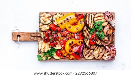 Grilled vegetables: red and yellow paprika, zucchini, eggplant, mushrooms, tomatoes and onions served on rustic wooden cutting board, white table background, top view Royalty-Free Stock Photo #2292366807